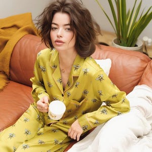 May include: A person wearing a yellow pajama set with a bee pattern, holding a white mug. The pajamas have a button-down top and matching pants. The person is sitting on a brown leather couch.
