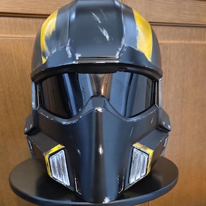 May include: A black and yellow helmet with a visor. The helmet has yellow stripes on top and yellow accents on the sides. The helmet is sitting on a black stand.