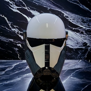Puede incluir: Un casco blanco y gris con una visera y detalles en negro. El casco tiene una parte superior redondeada y un diseño frontal detallado. La estética general sugiere un tema futurista o de ciencia ficción, posiblemente un disfraz o artículo de colección.