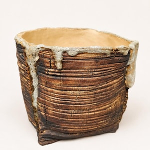 Puede incluir: Una maceta cilíndrica de cerámica con un exterior de color marrón oscuro profundamente texturizado que recuerda a la madera carbonizada. El borde presenta un esmalte gris claro grueso e irregular que gotea por el lateral.