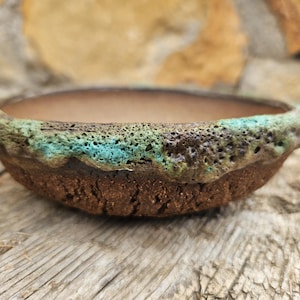 Può includere: Una piccola ciotola rotonda in ceramica con esterno marrone strutturato e bordo turchese e verde. L'interno è di colore marrone caldo. La ciotola viene probabilmente utilizzata per piante o scopi decorativi.