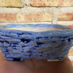 Puede incluir: Una maceta rectangular de cerámica para bonsái con una superficie exterior texturizada e irregular. El esmalte presenta una mezcla de tonos azul marino profundo y azul bígaro más claro, con un acabado interior liso blanquecino y pies.