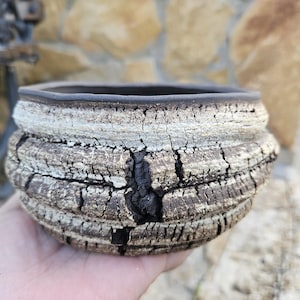 Könnte beinhalten: Handgefertigter, runder Keramik-Pflanztopf mit strukturierter, rissiger Oberfläche. Der Pflanztopf hat einen dunkelbraunen Innenraum und Rand, mit einer hellbraunen und grauen Außenseite. Das Design ähnelt gealtertem Holz.