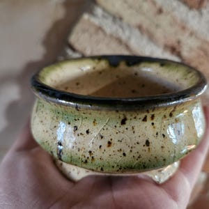 Può includere: Una piccola ciotola in ceramica fatta a mano con una glassa screziata verde chiaro e beige. Il bordo è marrone scuro e la ciotola poggia su tre piedini. Questa ciotola decorativa è adatta a piccole piante o come elemento decorativo unico.