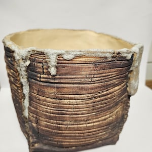 Puede incluir: Una maceta cuadrada de cerámica marrón texturizada con interior beige claro. La maceta tiene un aspecto rugoso y en capas, con un material más claro y espumoso alrededor del borde. El exterior tiene un veteado similar a la madera.