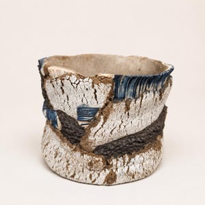 Puede incluir: Un recipiente cilíndrico de cerámica con una superficie exterior blanca, agrietada y muy texturizada. El diseño incorpora bandas contrastantes de color marrón oscuro con textura rugosa y acentos de esmalte azul profundo.