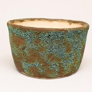 Peut inclure: Un petit bol en céramique rond avec un extérieur texturé et poreux présentant un motif tacheté de brun terreux et de vert turquoise vif. L'intérieur du bol est lisse et fini avec un émail uni de couleur crème claire.