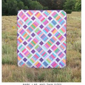 May include: A colorful quilt pattern called "The Iris Quilt" by Erica Jackman. The quilt is made with a variety of bright colors and patterns, including pink, blue, green, and yellow. The quilt is displayed on a grassy field.