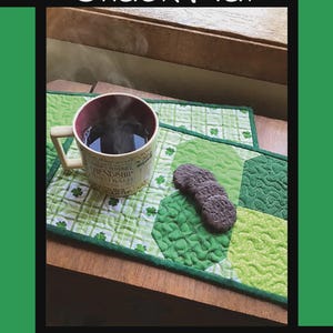 May include: A rectangular quilted snack mat featuring various shades of green fabric with shamrock patterns. A ceramic mug with steam and three dark round cookies are placed on the mat. The text reads Shamrock Snack Mat, Tricia Lynn Maloney, Orphan Quilt Designs, for Villa Rosa Designs.