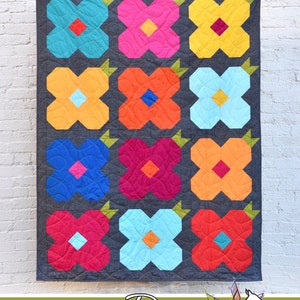 May include: A quilt block pattern called "May Flowers" with a floral design in bright colors like blue, orange, pink, and yellow. The quilt block is on a gray background. The quilt block is made by Running Doe Quilts for Villa Rosa Designs.