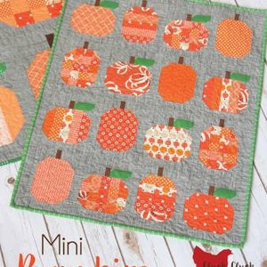May include: A gray fabric quilt with a green border features a pattern of 16 orange and white mini pumpkins. The quilt is titled "Mini Pumpkins" and has the text "Cluck Cluck Sew" in the bottom right corner.