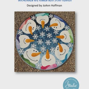 May include: A blue and white fabric quilt with a circle design featuring eight snowmen faces with orange noses and black eyes. The quilt is surrounded by white snowflakes. The text "Kisses from Heaven" and "Snowflakes are kisses sent from Heaven" is at the top of the image. The text "Designed by JoAnn Hoffman" is below the quilt.