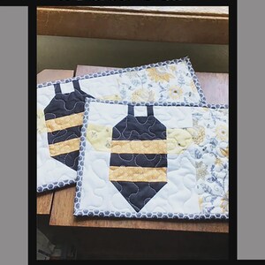 May include: Two rectangular quilted snack mats featuring a central bee motif with black and yellow stripes. The mats have a white background with a floral pattern on the side and a grey honeycomb border. Text reads Buzzy Bee Snack Mat, Tricia Lynn Maloney, Orphan Quilt Designs, for Villa Rosa Designs.