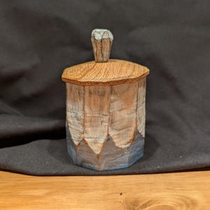 May include: A hand-carved wooden box with a lid and a small handle. The box is cylindrical with a textured, light-coloured exterior and a darker wooden lid. The handle on the lid is a lighter colour. The box sits on a wooden surface.