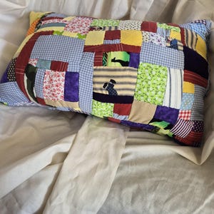 以下が含まれることがあります： 花柄、ギンガムチェック、ストライプなど、さまざまな生地の模様と色を使用した長方形のパッチワーク枕。枕は柔らかく、質感のある外観で、ふわふわの素材が詰められています。枕は白いシーツの上に置かれています。