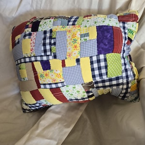 May include: A patchwork pillow with a variety of fabric patterns and colors. The pillow features squares of yellow, blue gingham, floral, and solid fabrics. The pillow is rectangular and appears to be handmade.