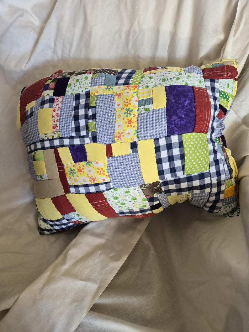 以下が含まれることがあります： 様々な柄と色の生地を使った長方形のパッチワーク枕。黄色、青いギンガムチェック、花柄、無地の生地の正方形が特徴です。手作りのように見えます。