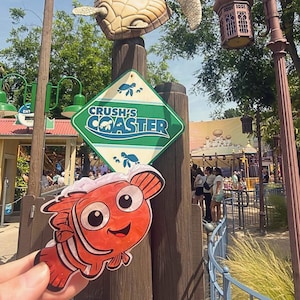 Peut inclure: Une petite découpe plate de poisson-clown orange et blanc tenue au premier plan, devant un poteau en bois portant un panneau en losange indiquant Crush's Coaster, surmonté d'une grande figurine sculptée de tortue de mer.