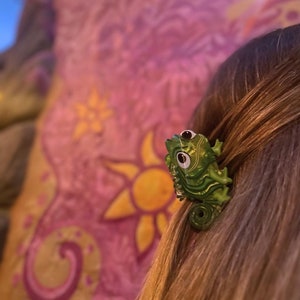 May include: A green chameleon hair clip with large eyes. The clip is made of plastic and has a detailed design.