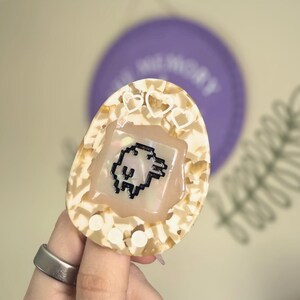 May include: A cream and tan retro handheld game device featuring a pixelated character. The device has a heart-shaped design and is held by a person wearing a silver ring. A purple disc is visible in the background.