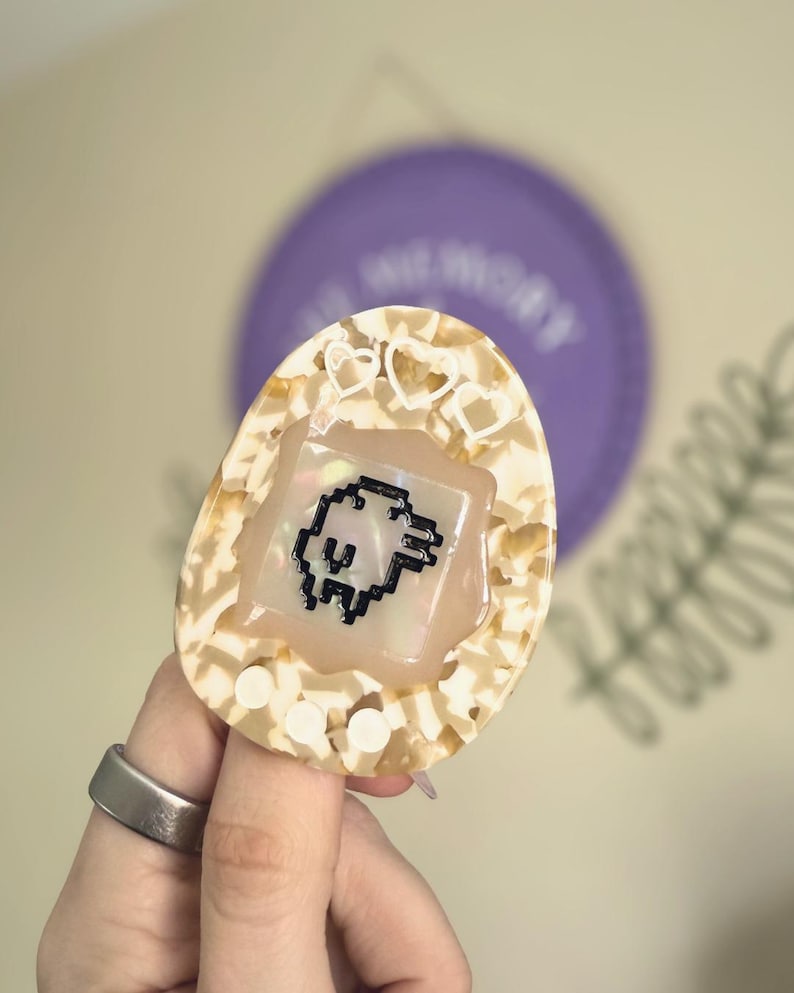 May include: A cream and tan retro handheld game device featuring a pixelated character. The device has a heart-shaped design and is held by a person wearing a silver ring. A purple disc is visible in the background.