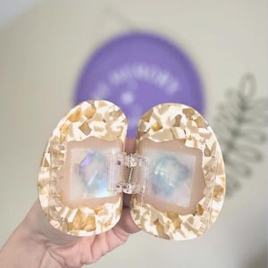 May include: Open, oval-shaped hair clip with a speckled, cream and tan pattern. The clip has a clear, rectangular centre with iridescent accents and a silver-coloured spring hinge. The background includes a purple balloon.