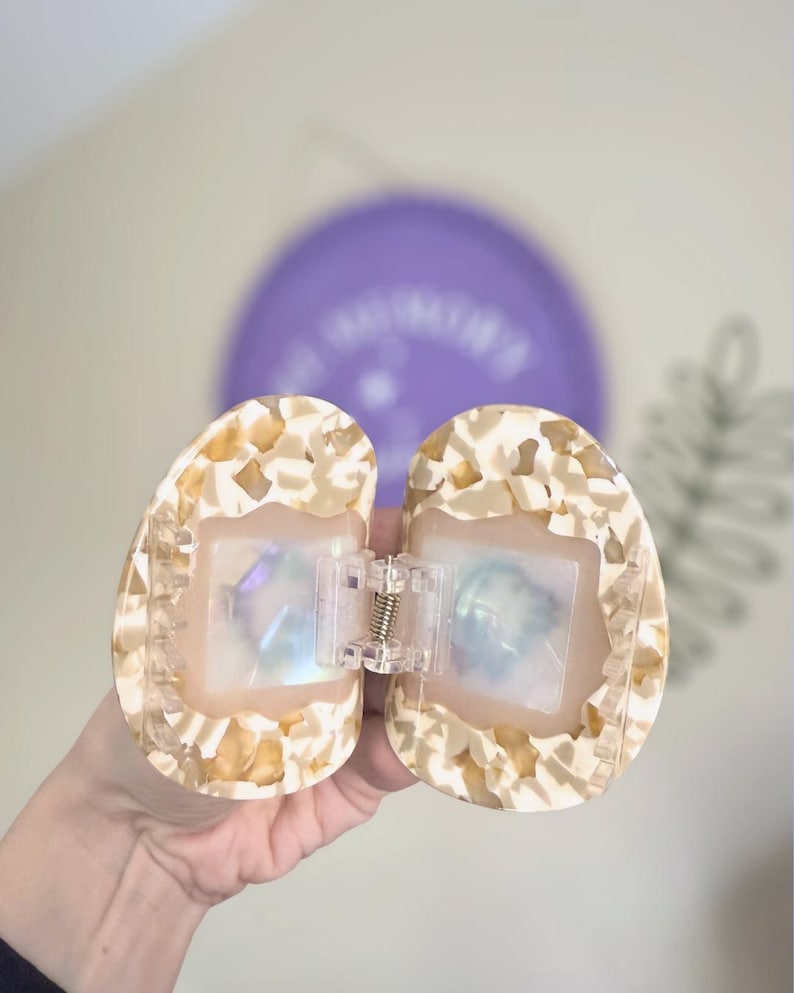 May include: Open, oval-shaped hair clip with a speckled, cream and tan pattern. The clip has a clear, rectangular centre with iridescent accents and a silver-coloured spring hinge. The background includes a purple balloon.