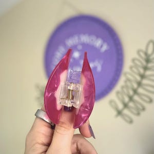 May include: A pink and clear hair claw clip with a gold-coloured spring mechanism. The clip is held open, revealing the inner workings. The background includes a purple circle with white text and a green plant.