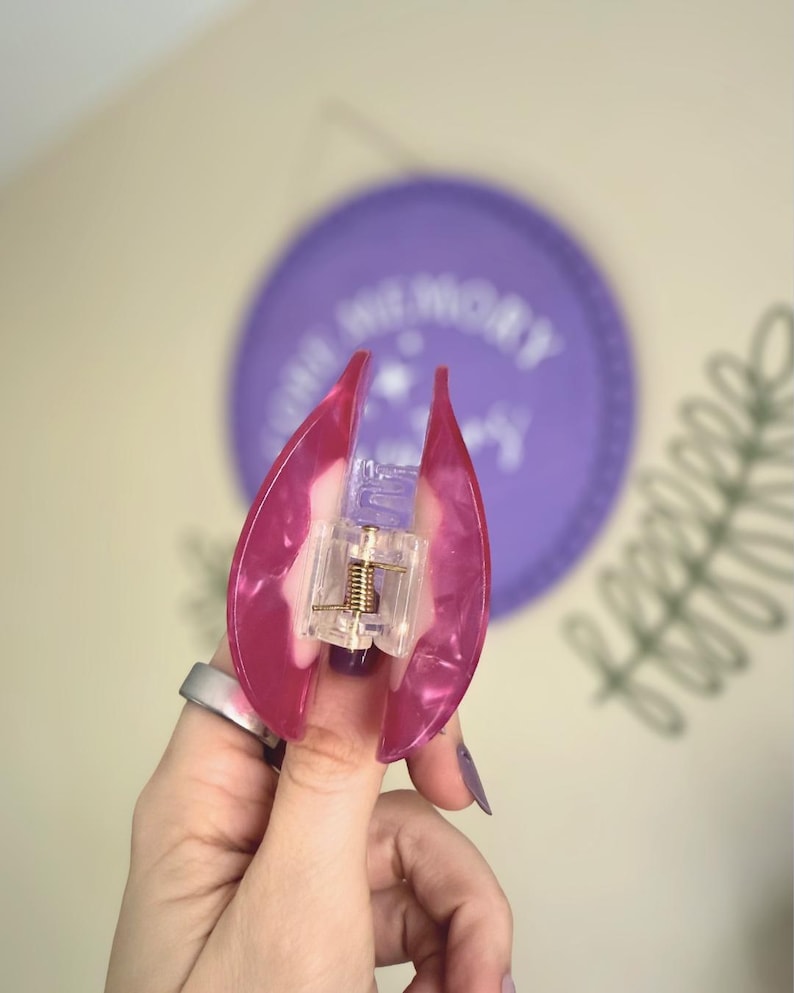 May include: A pink and clear hair claw clip with a gold-coloured spring mechanism. The clip is held open, revealing the inner workings. The background includes a purple circle with white text and a green plant.