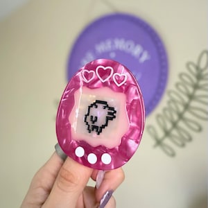 May include: A pink and white marbled-effect accessory shaped like a retro handheld game. The game has a pixelated animal graphic, three white hearts, and three white dots. A purple disc with the word "MEMORY" is in the background.