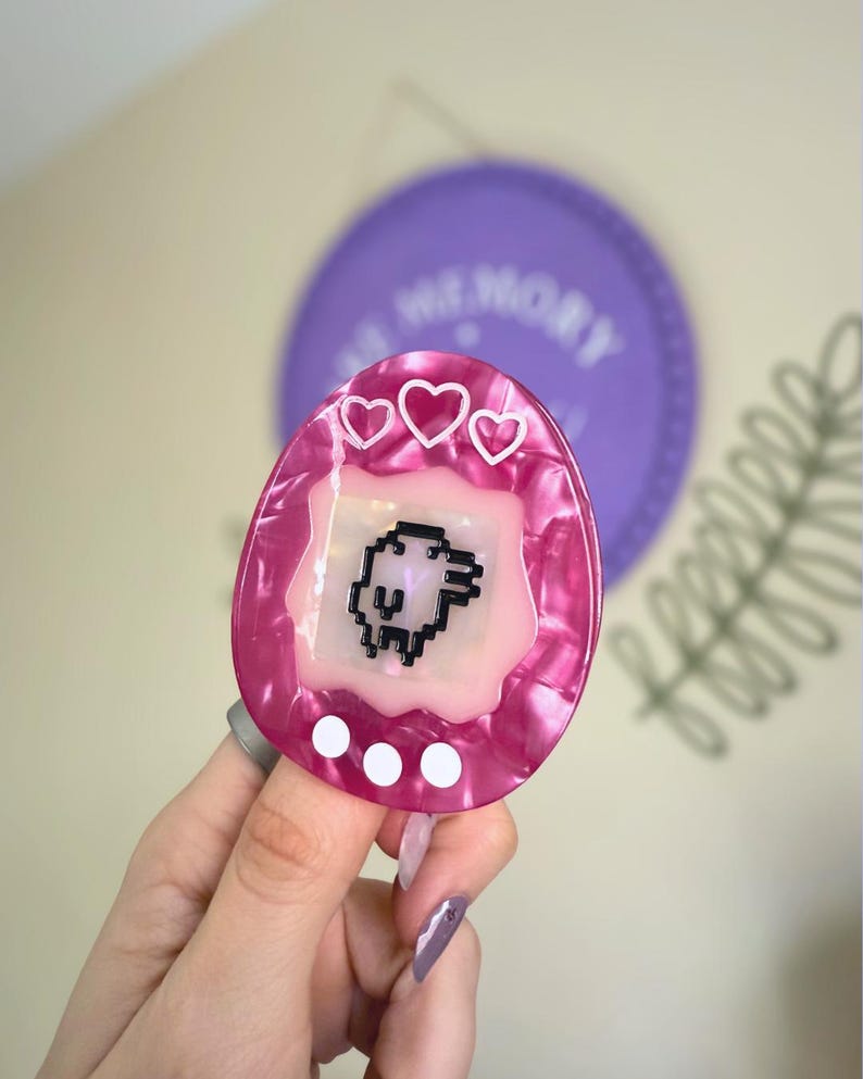 May include: A pink and white marbled-effect accessory shaped like a retro handheld game. The game has a pixelated animal graphic, three white hearts, and three white dots. A purple disc with the word "MEMORY" is in the background.