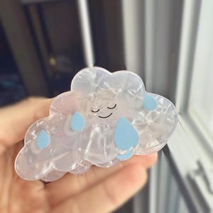 Op de afbeelding: Een haarclip in de vorm van een wolk met een lachend gezicht en blauwe regendruppelaccenten. De clip is gemaakt van een parelmoerachtig, doorschijnend materiaal. Het haaraccessoire is ontworpen om het haar op zijn plaats te houden.