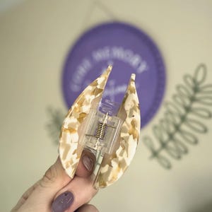 May include: A tortoiseshell patterned hair claw clip in cream and tan hues. The clip features a clear plastic mechanism with a metal spring. A purple circle with text and a green leaf design are in the background.