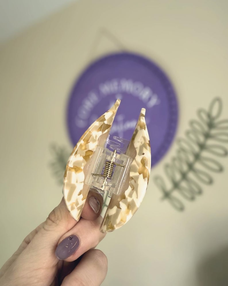 May include: A tortoiseshell patterned hair claw clip in cream and tan hues. The clip features a clear plastic mechanism with a metal spring. A purple circle with text and a green leaf design are in the background.