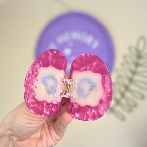 May include: A pink hair claw clip with a heart pattern. The clip is made of translucent plastic and has a clear hinge with a gold-coloured spring. The clip is open, revealing a light-coloured interior.
