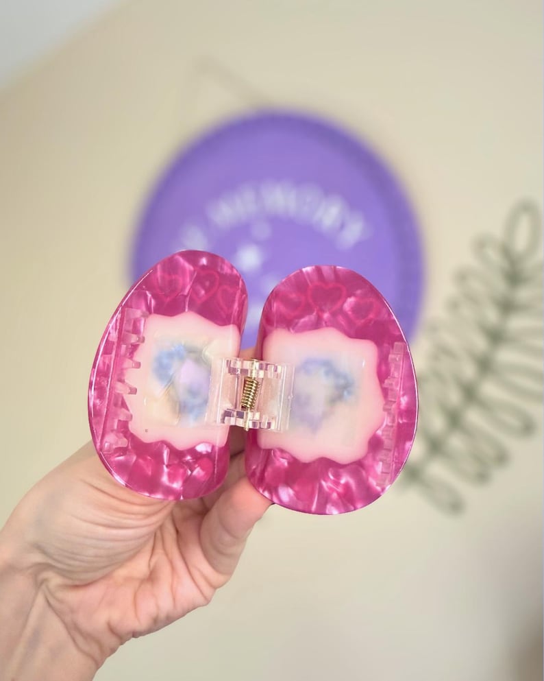 May include: A pink hair claw clip with a heart pattern. The clip is made of translucent plastic and has a clear hinge with a gold-coloured spring. The clip is open, revealing a light-coloured interior.