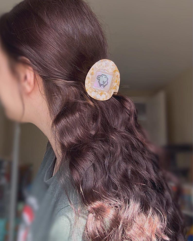 May include: An egg-shaped hair clip with a retro pixelated creature graphic. The clip has a pearlescent, speckled design in cream and gold. The clip is attached to wavy brown hair. A stylish hair accessory.