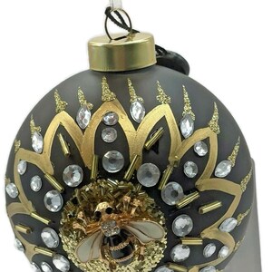 May include: A decorative Christmas ornament with a dark gray base, adorned with gold floral patterns, clear crystals, and a detailed bee emblem. The ornament has a gold cap and a white ribbon for hanging.