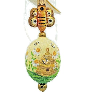 May include: A decorative ornament featuring a bee and a beehive design. The bee has gold glitter wings and a striped body. The egg-shaped ornament has a yellow background with a beehive, daisies, and bees. The bottom is green.