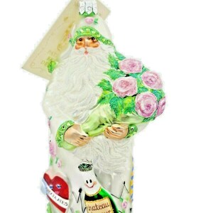 May include: A decorative Santa Claus ornament holding a bouquet of pink roses. The ornament is white with green accents and features a heart and a champagne bottle with the text "Chateau Amour".