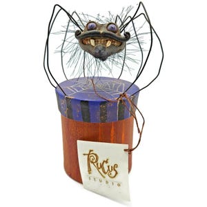 May include: A whimsical, handcrafted spider figurine with a smiling face, perched atop a wooden box. The box features a blue lid with a spiderweb design and a brown body. A tag reads "Rucus Studio".