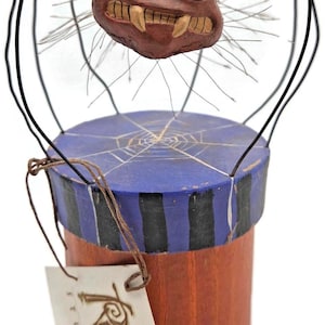 May include: A whimsical, handcrafted spider figurine with a brown, toothy face and large orange eyes. The spider sits atop a cylindrical box with a blue lid featuring a white spiderweb design. The box has a striped pattern and a wooden base.
