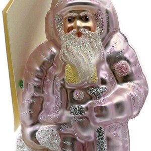 May include: A pink Christmas ornament shaped like Santa Claus, holding a sack with "2000" on it and a silver spoon. The ornament has glitter accents and a gold-colored beard. A tag is attached to the ornament.