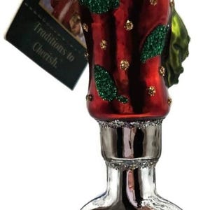 May include: A Christmas ornament shaped like a bottle, with a red body adorned with green glitter leaves and gold accents. The bottom is silver with blue glitter. A tag reads "Traditions to Cherish."