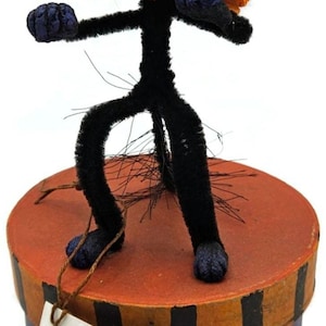May include: A whimsical Halloween cat figure with a black body and a head with blue accents and orange ruff. The figure stands on a round box with orange and black stripes. The tag reads "Rucus Studio".
