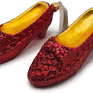 May include: A pair of red glittered shoes with a yellow interior. The shoes have a small heel and a decorative strap across the top. The shoes are likely ornaments or decorative items.