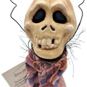 May include: A decorative skull ornament with a wire halo. The skull is beige with black eye sockets and a toothy grin. It wears a pink and blue striped scarf. A tag is attached.