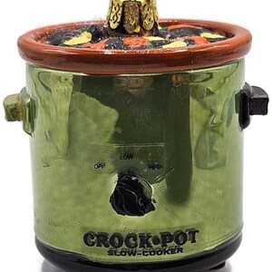 May include: A green glass Crock-Pot slow-cooker Christmas ornament. The ornament has a brown rim and a gold-colored top. The text "CROCK-POT SLOW-COOKER" is printed on the front. The ornament has a black knob with "OFF", "LOW", and "HIGH" settings.