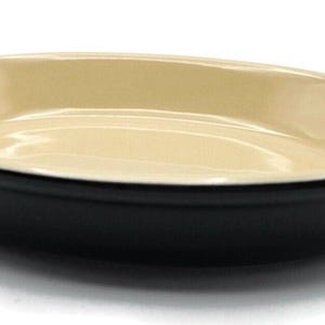 May include: An oval-shaped, black and cream-colored ceramic baking dish. The dish has a black exterior, a cream-colored interior, and two handles. The dish is designed for baking and serving food.