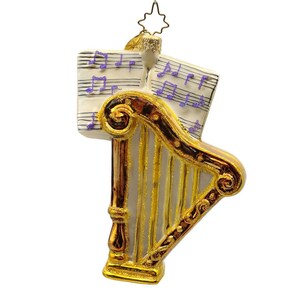 May include: A decorative Christmas ornament featuring a golden harp with a brown trim. Two sheets of music with purple notes are behind the harp. The ornament is suspended by a thin wire and a star-shaped hanger.